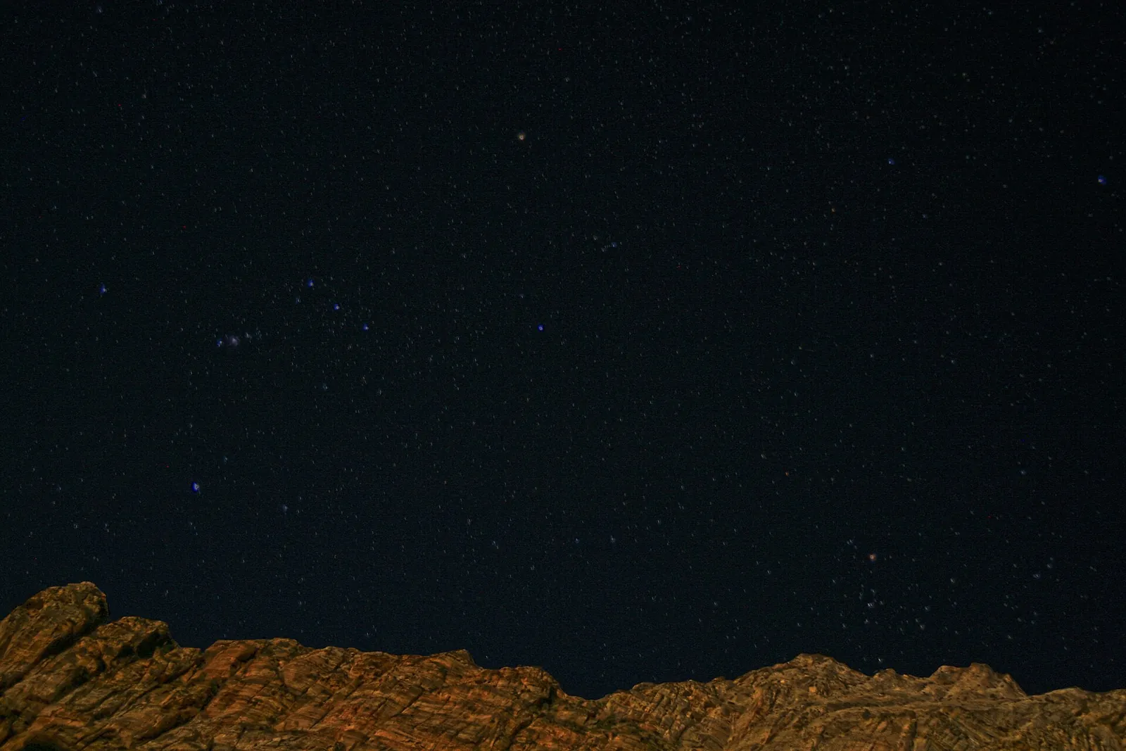 Stars of the constellation Orion in a dark sky above a faintly lit cliff line in Zion National Park.