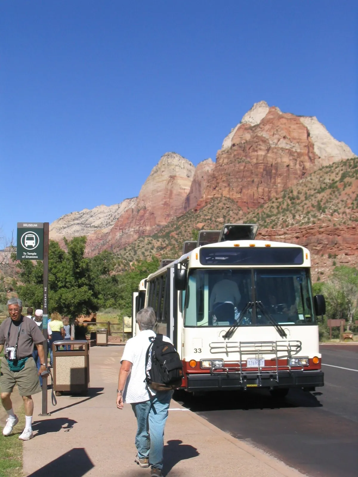 A park shuttle bus at the Zion Human History Museum stop with red peaks behind it.
